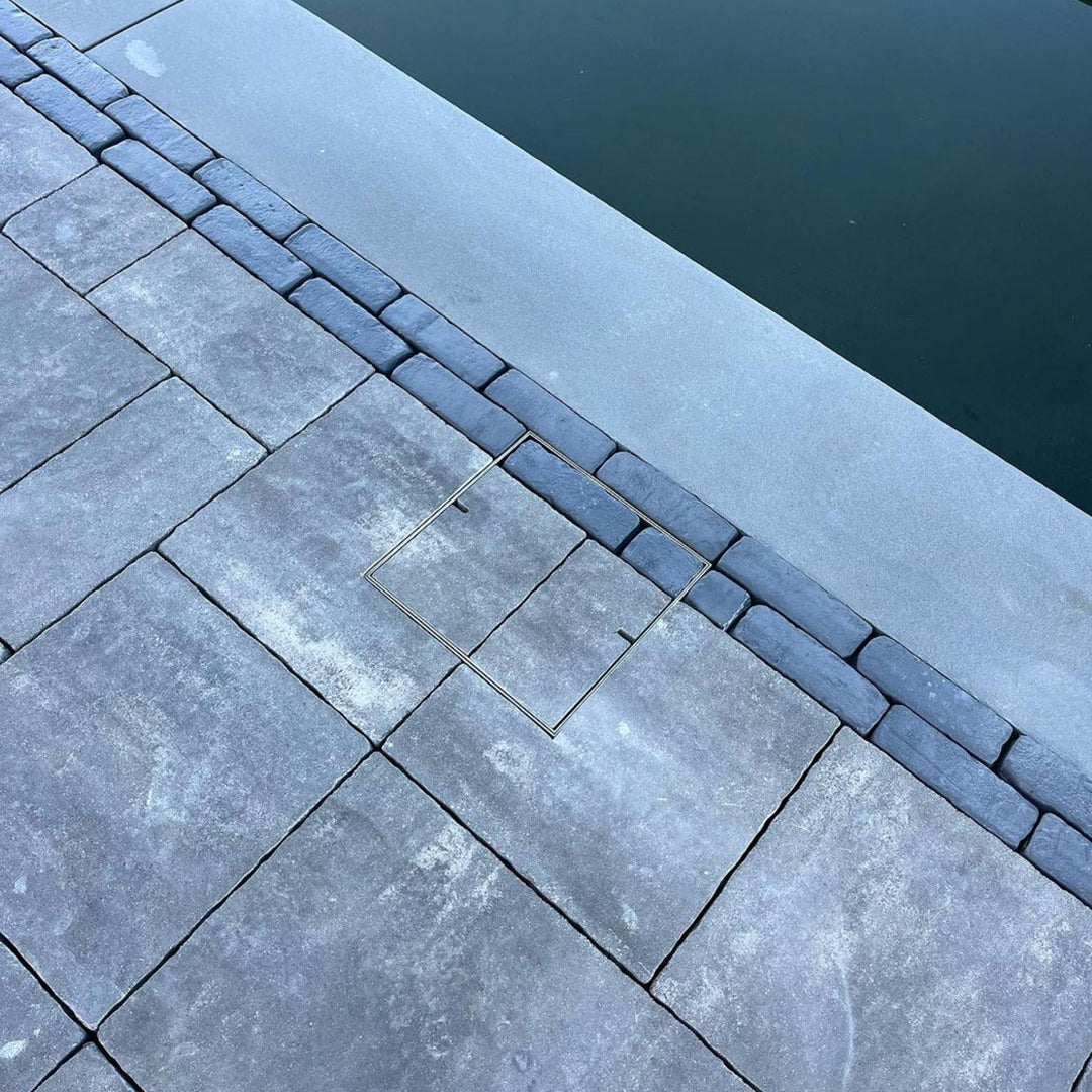 Close-up of grey stone pavers beside a pool, showing a flush-fitting square HIDE Skimmer Cover set into the paving near the water’s edge.