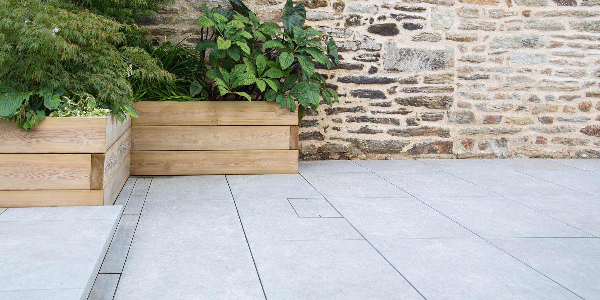 Flush HIDE stainless steel access cover integrated into large-format pavers beside timber planters and stone wall.