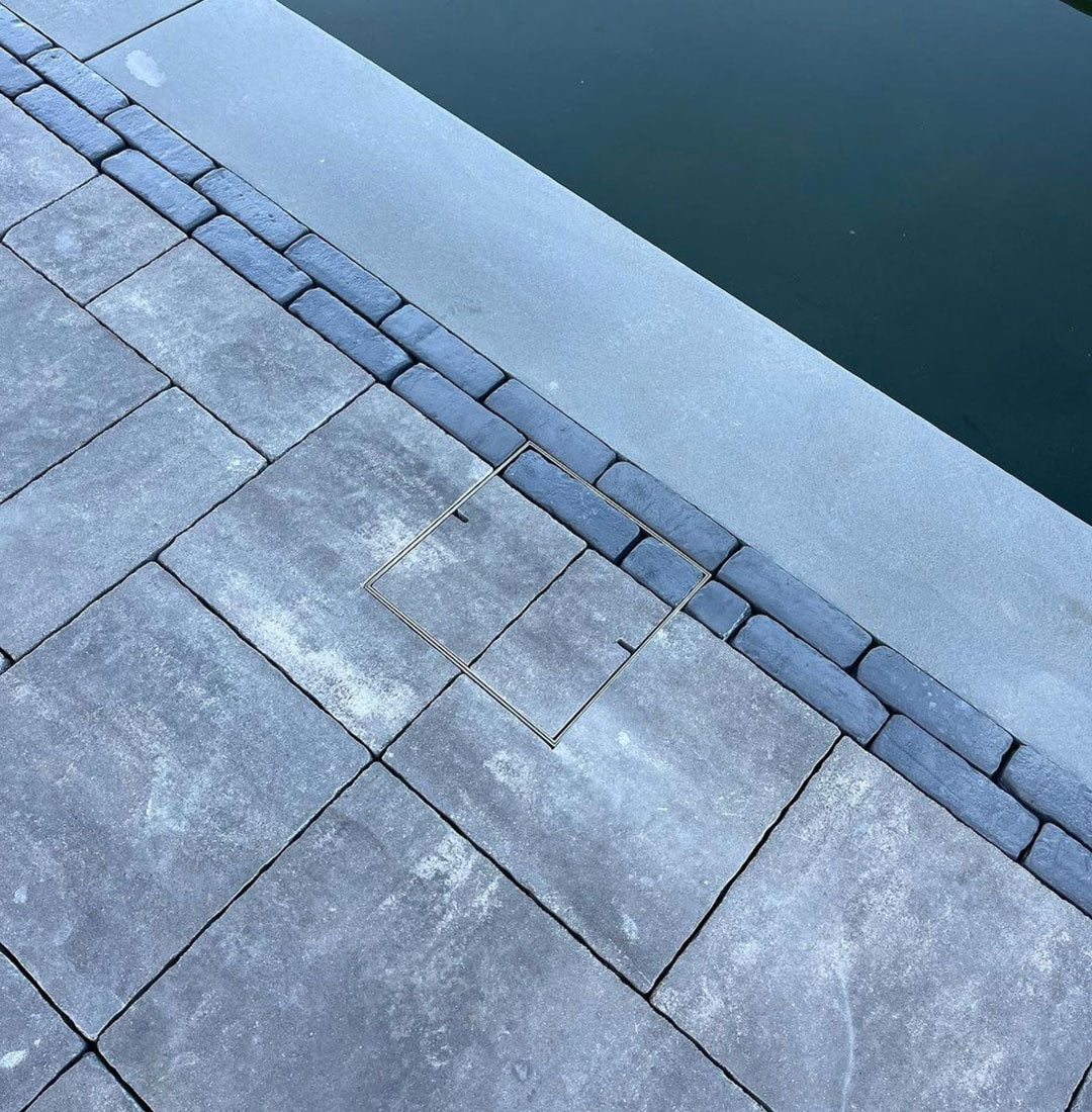 Close-up of grey stone pavers beside a pool, showing a flush-fitting square HIDE Skimmer Cover set into the paving near the water’s edge.
